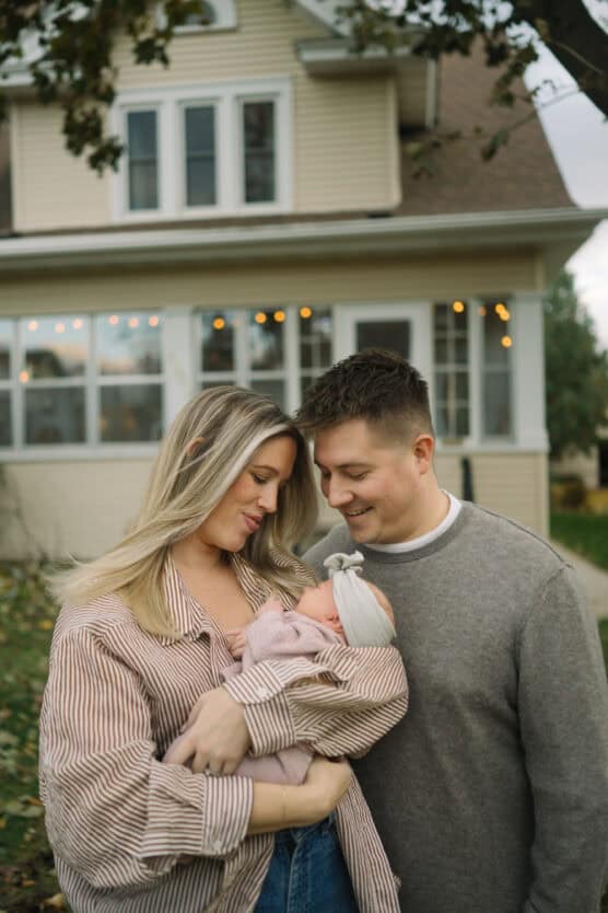 A couple stands close together outside a house, smiling down at a baby wrapped in a pink blanket and wearing a light headband, who is cradled in the woman’s arms. by Justin Salem Meyer
