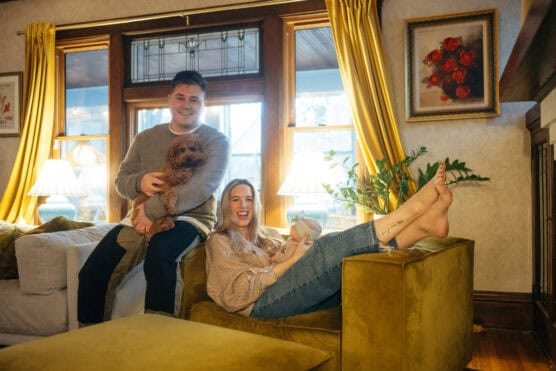 A smiling man holds a brown dog while standing behind a woman lounging barefoot on a yellow couch, holding a baby. They are in a warmly lit living room with yellow curtains and framed art on the walls. by Justin Salem Meyer