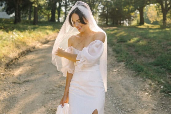 A bride in an off-the-shoulder white dress and veil smiles while standing on a sunlit dirt path surrounded by greenery and trees. by Justin Salem Meyer