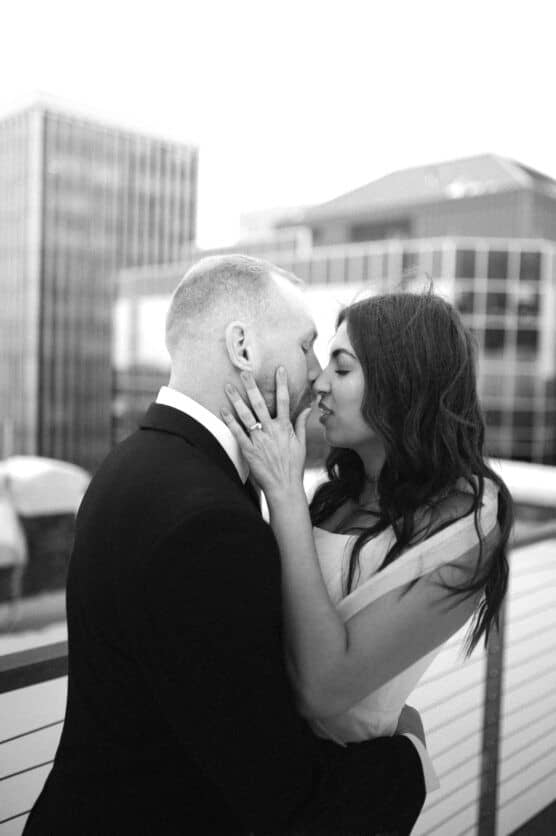 A couple in formal attire shares an intimate moment on a rooftop, with city buildings in the background. The woman is gently holding the mans face as they lean in for a kiss. The photo is in black and white. by Justin Salem Meyer