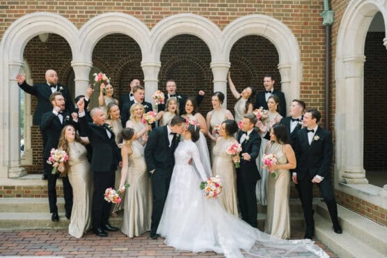 A bride and groom stand together, surrounded by their wedding party in formal attire. The bridesmaids wear gold dresses and hold pink bouquets; the groomsmen wear black tuxedos. They all celebrate in front of an arched brick building. by Justin Salem Meyer