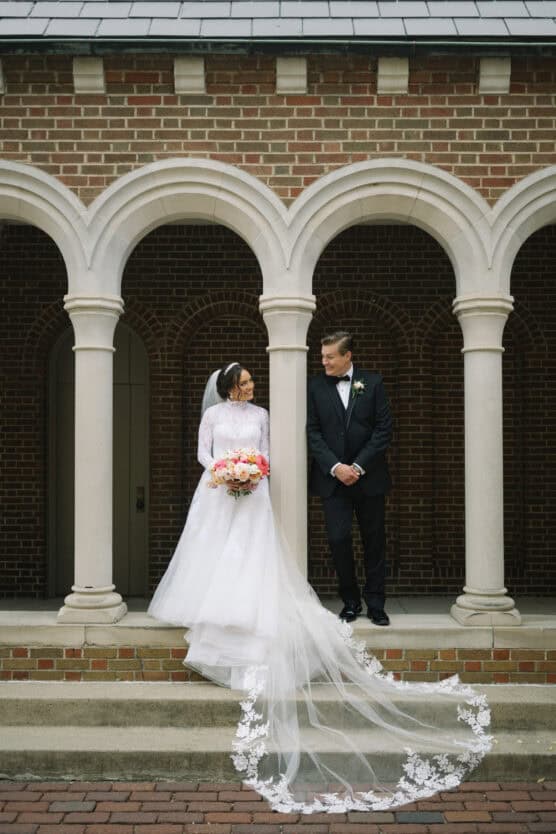 A bride in a white gown and veil holding a bouquet stands beside a groom in a black tuxedo under stone arches of a brick building, both smiling at each other. The bride’s long lace train cascades down the steps. by Justin Salem Meyer