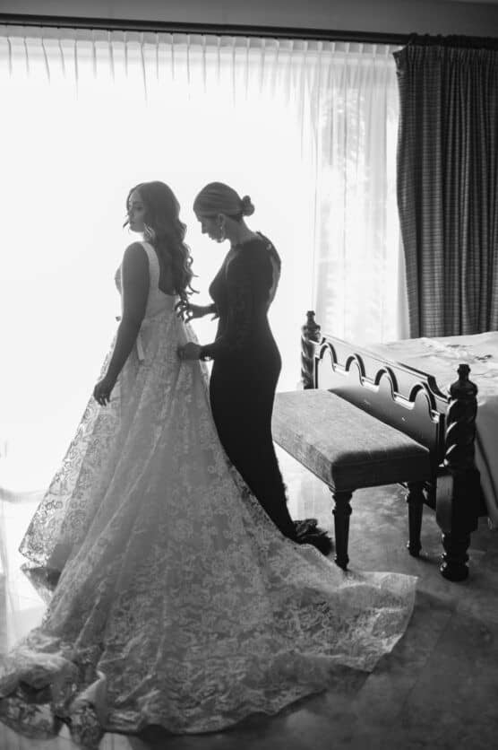 A woman in a lace wedding gown stands while another woman helps adjust her dress in a softly lit room with large windows, heavy curtains, and a bench near the bed. by Justin Salem Meyer