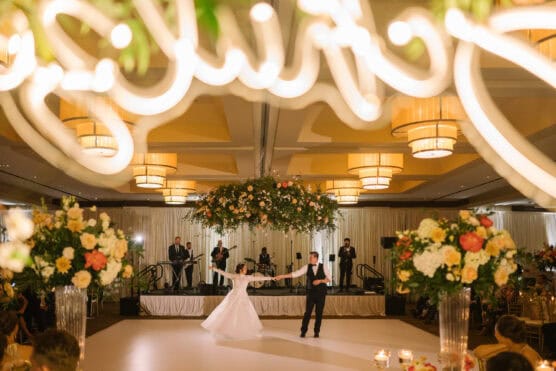 A bride and groom dance together in a ballroom decorated with flowers and hanging lights, while a live band performs on stage in the background. Elegant floral arrangements frame the scene. by Justin Salem Meyer