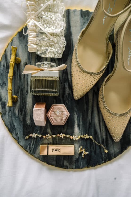 Elegant flat lay of wedding accessories: beige rhinestone heels, lace garter, perfume bottle, lipstick, gold bracelet, necklace, and an engagement ring in a pink hexagonal box, all arranged on a dark marble tray. by Justin Salem Meyer