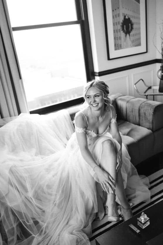 A smiling bride in a flowing wedding gown sits on a sofa, adjusting her shoe. Light streams through a window behind her, and a framed photo hangs on the wall. The scene is elegant and serene in black and white. by Justin Salem Meyer
