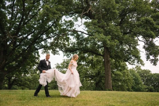A groom in a black tuxedo helps a bride in a white wedding dress walk across a grassy field, surrounded by large green trees under a cloudy sky. by Justin Salem Meyer