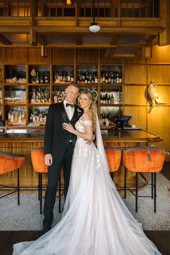 A bride in a flowing white gown and a groom in a black tuxedo pose together, smiling, in front of a wooden bar with shelves of bottles and orange chairs. by Justin Salem Meyer
