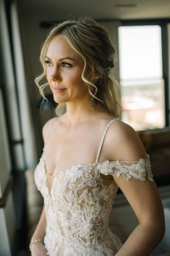 A woman in a lace, off-shoulder wedding dress stands by a window, looking outside with a soft smile. Her blonde hair is styled in loose waves, and natural light highlights her face. by Justin Salem Meyer