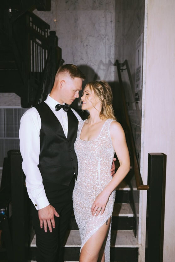 A couple dressed formally smiles and looks into each others eyes on a staircase. The man wears a black vest and bow tie, while the woman wears a sparkling, sleeveless gown with a thigh-high slit. by Justin Salem Meyer