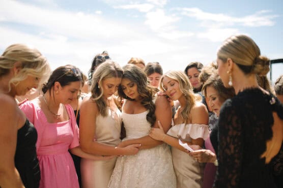 A bride stands with her eyes closed, surrounded by bridesmaids and loved ones in soft pastel dresses, sharing an emotional, supportive moment together under a bright, partly cloudy sky. by Justin Salem Meyer