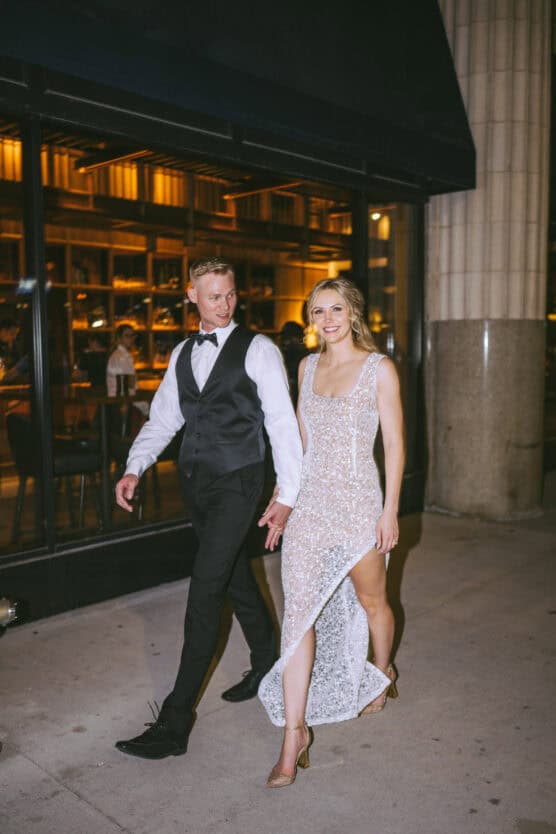 A man in a black tuxedo and a woman in a sparkly, sleeveless dress with a thigh-high slit are walking together on a city sidewalk at night, smiling and holding hands. by Justin Salem Meyer