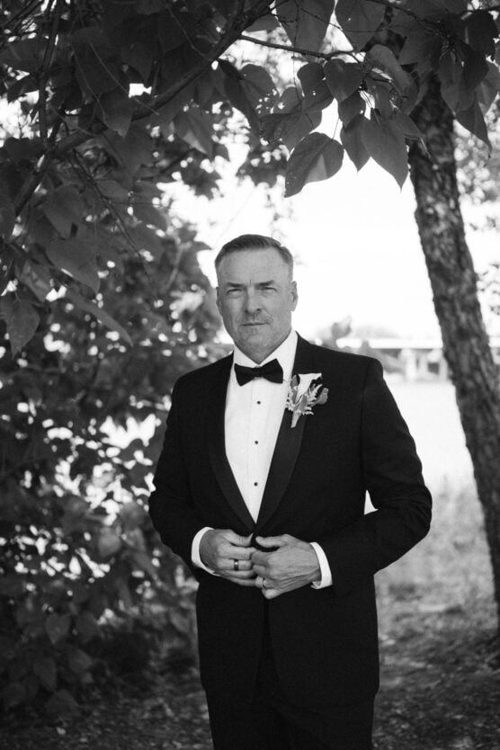 A man in a tuxedo stands outdoors under leafy branches, looking at the camera with a serious expression. He wears a bow tie and boutonniere, and holds his hands together in front of him. The photo is in black and white. by Justin Salem Meyer