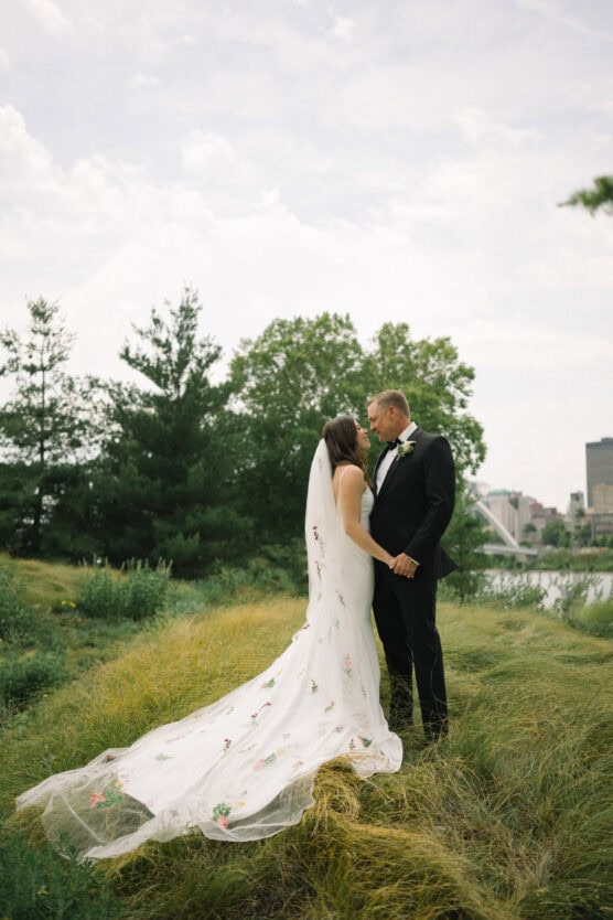 A bride in a white floral gown and veil stands facing a groom in a black tuxedo on a grassy field, holding hands and gazing at each other, with trees and a city skyline in the background. by Justin Salem Meyer