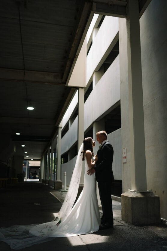 A bride in a white dress and veil stands closely with a groom in a dark suit, embracing under a beam of sunlight in a shadowy urban parking garage. by Justin Salem Meyer