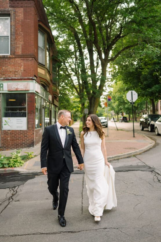 A bride in a white dress and a groom in a black tuxedo walk hand in hand on a city street, smiling at each other, with trees and brick buildings in the background. by Justin Salem Meyer
