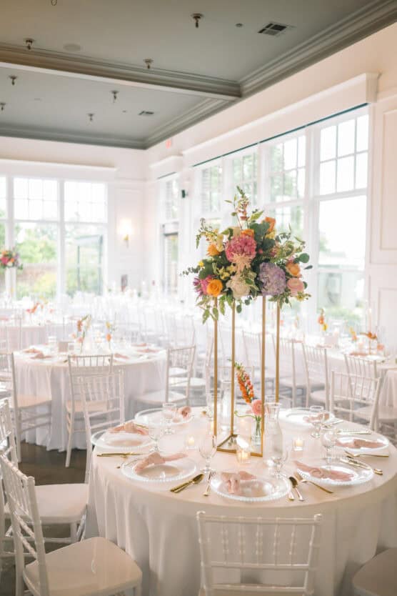 A round table set for a formal event with white linens, clear plates, gold cutlery, and white chairs. A tall floral centerpiece with colorful flowers decorates the table. Sunlight streams in through large windows. by Justin Salem Meyer