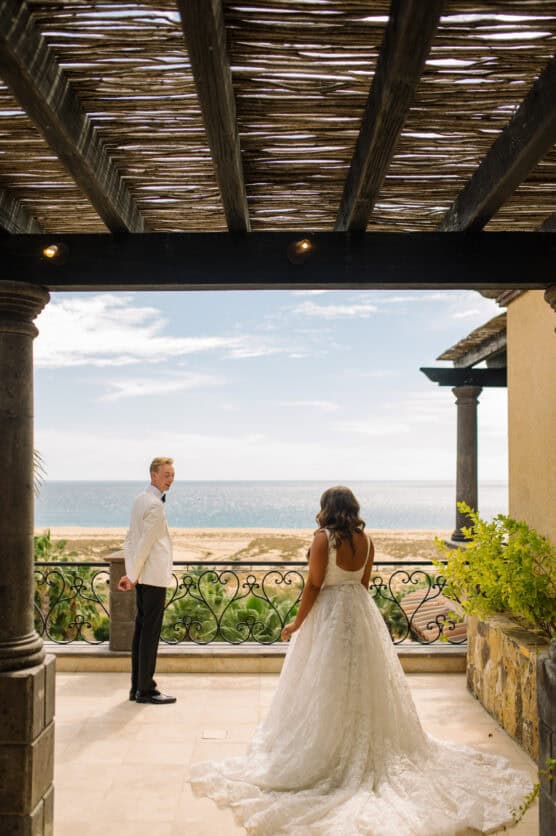 A bride in a white gown approaches a groom in a white jacket on a terrace overlooking a beach and ocean, framed by stone columns and wooden pergola. by Justin Salem Meyer