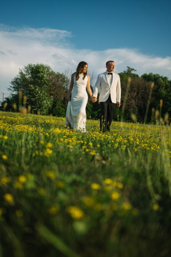 A couple in formal attire walks hand in hand through a field of yellow wildflowers under a blue sky, with greenery and trees in the background. by Justin Salem Meyer