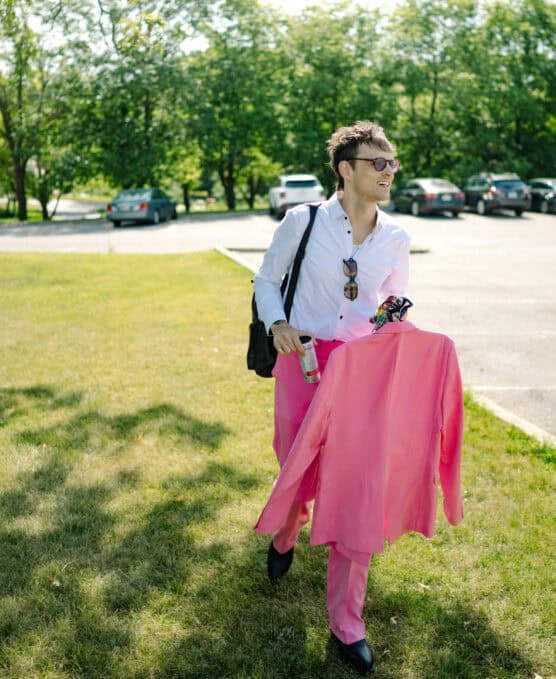 A man in a white shirt and pink pants carries a matching pink jacket, sunglasses, and a drink can while walking on grass in a sunny parking lot with trees and parked cars in the background. by Justin Salem Meyer