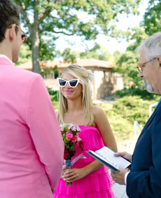 A woman in a bright pink dress and white sunglasses holds a bouquet and smiles during an outdoor ceremony. A man in a pink suit faces her, while an officiant, holding a notebook, stands beside them. Trees and buildings are in the background. by Justin Salem Meyer
