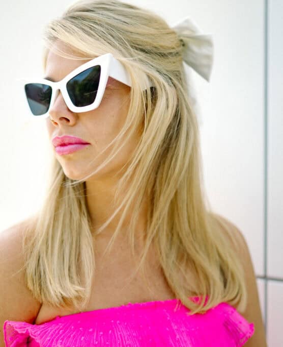 A woman with long blonde hair, wearing a large white hair bow, white cat-eye sunglasses, and a bright pink strapless top, looks to the side against a light background. by Justin Salem Meyer