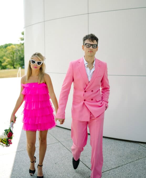 A woman in a bright pink ruffled dress and white sunglasses holds a bouquet and walks hand in hand with a man in a matching pink suit and black glasses. They are outside near a modern white building. by Justin Salem Meyer