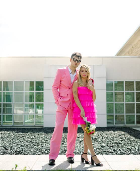 A person in a bright pink suit stands beside another in a ruffled hot pink dress holding flowers. They pose outside a modern building with large windows, both looking at the camera. by Justin Salem Meyer