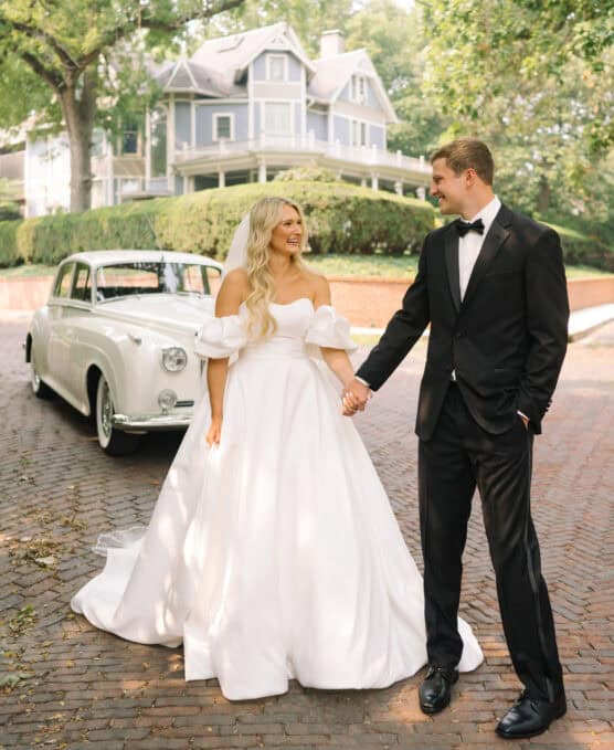 A bride in a white gown and a groom in a black tuxedo hold hands and smile at each other on a cobblestone path, with a classic white car and a large house with a porch and trees in the background. by Justin Salem Meyer