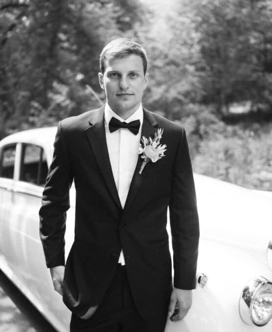A man in a tuxedo with a bow tie and boutonniere stands in front of a classic car outdoors, looking at the camera. The photo is in black and white. by Justin Salem Meyer