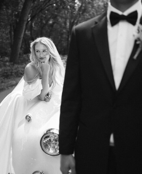 A bride in a white dress leans on the hood of a vintage car, looking thoughtfully to the side, while a groom in a tuxedo stands in the foreground, partially out of focus. The scene is outdoors with trees in the background. by Justin Salem Meyer