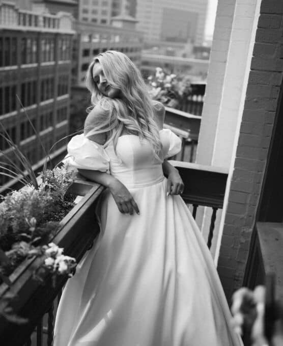 A woman in a long, elegant off-shoulder wedding dress stands on a balcony, leaning against the railing with flowers in the planter. City buildings are visible in the blurred background. The image is in black and white. by Justin Salem Meyer
