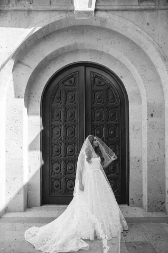 A bride in a lace wedding gown and veil stands in front of a large ornate arched wooden door, posing gracefully. The scene is lit with natural sunlight, casting distinct shadows on the stone wall. by Justin Salem Meyer
