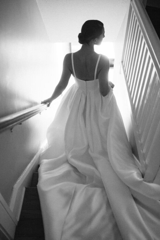 A woman in a flowing white wedding gown descends a staircase, holding the rail and her dress, with light streaming in from above. The image is captured in black and white. by Justin Salem Meyer