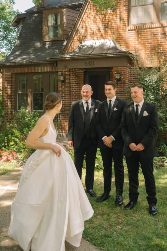 A bride in a white dress walks toward three men in black suits and ties, who are smiling at her, outside a brick house on a sunny day. by Justin Salem Meyer
