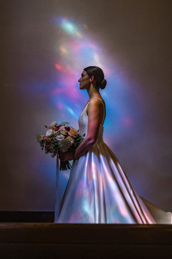 A bride in a white gown holding a bouquet stands in profile against a plain wall, illuminated by colorful, soft light reflections that create a dreamy, ethereal effect on her dress and the background. by Justin Salem Meyer
