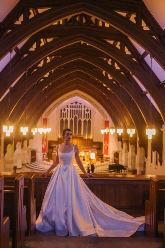 A bride in a white satin gown with a long train stands inside a grand, warmly lit church with high vaulted wooden arches and glowing chandeliers. by Justin Salem Meyer