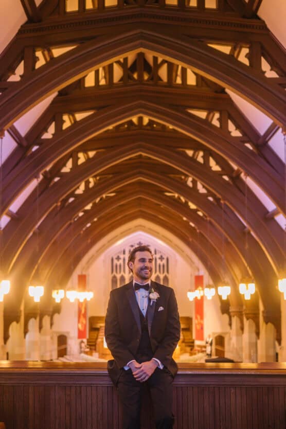 A man in a black tuxedo and bow tie stands smiling in a church with high arched wooden ceilings and warm hanging lights, creating a dramatic, elegant atmosphere. by Justin Salem Meyer