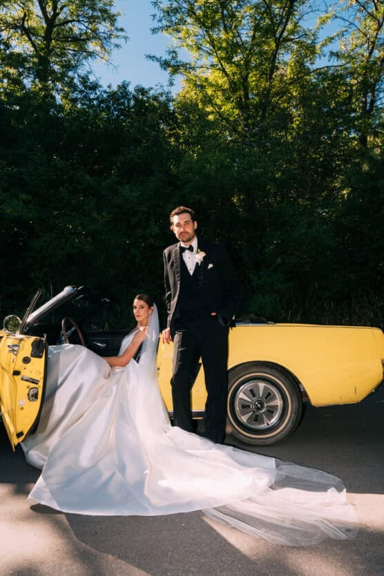 A bride in a white gown sits in a yellow convertible while the groom in a black tuxedo stands beside her. They are outdoors, with green trees and sunlight in the background. by Justin Salem Meyer