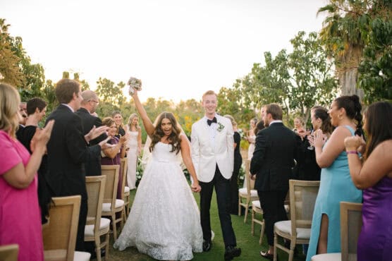 A bride in a white gown raises her bouquet triumphantly while holding hands with the groom in a white tuxedo jacket as they walk down the aisle, surrounded by applauding guests outdoors. by Justin Salem Meyer