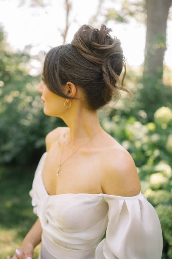 A woman in an off-the-shoulder white dress with sheer sleeves stands outdoors, facing sideways. Her brown hair is styled in an elegant updo, and she wears a gold necklace and hoop earrings. Greenery is blurred in the background. by Justin Salem Meyer