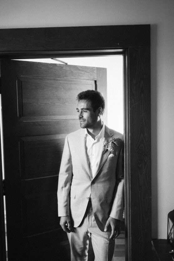 A man in a light-colored suit with a boutonnière stands in a doorway, looking to the side and smiling. The photo is in black and white. by Justin Salem Meyer