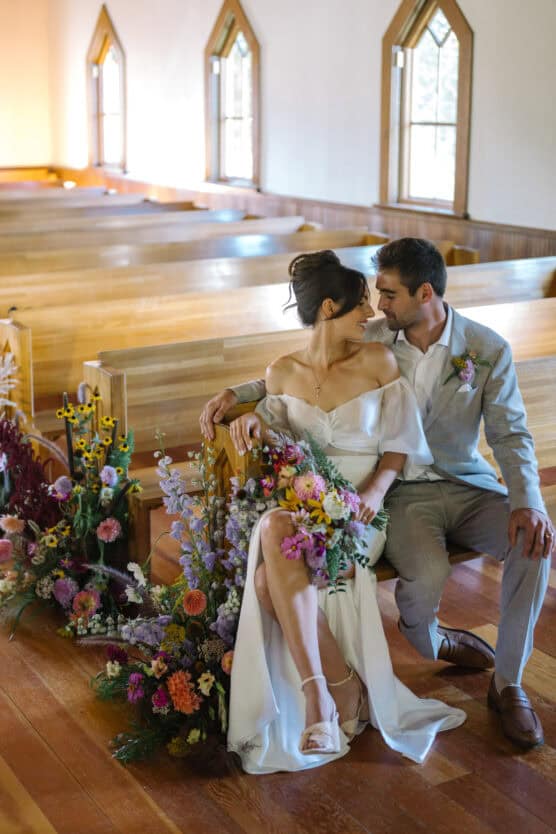 A bride in an off-shoulder white dress and a groom in a light gray suit sit close together in a sunlit church, surrounded by colorful flowers and wooden pews, looking lovingly into each others eyes. by Justin Salem Meyer