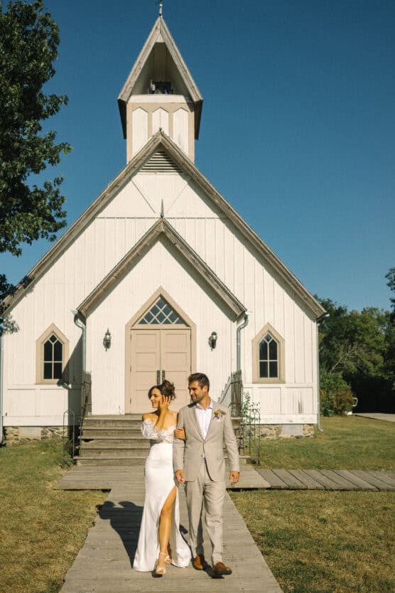 A bride in a white dress and a groom in a light suit walk hand in hand outside a small white wooden church on a sunny day, with clear blue sky in the background. by Justin Salem Meyer