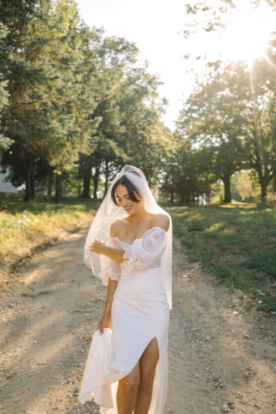 A bride in an off-the-shoulder white dress and veil stands on a sunlit dirt path, surrounded by trees and greenery, smiling and gently holding her dress. by Justin Salem Meyer