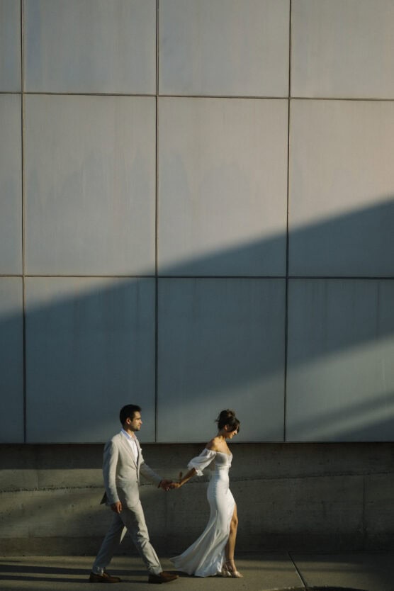 A bride in a white dress and a groom in a light suit walk hand in hand along a sidewalk, casting long shadows against a large, modern tiled wall. by Justin Salem Meyer