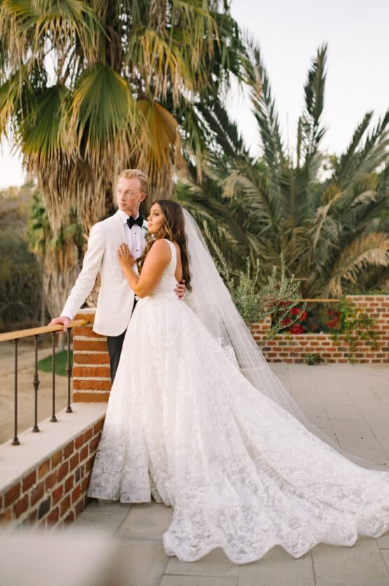 A bride in a white lace gown and veil stands beside a groom in a white tuxedo jacket and black bow tie. They pose outdoors by a brick railing, surrounded by palm trees and greenery. by Justin Salem Meyer