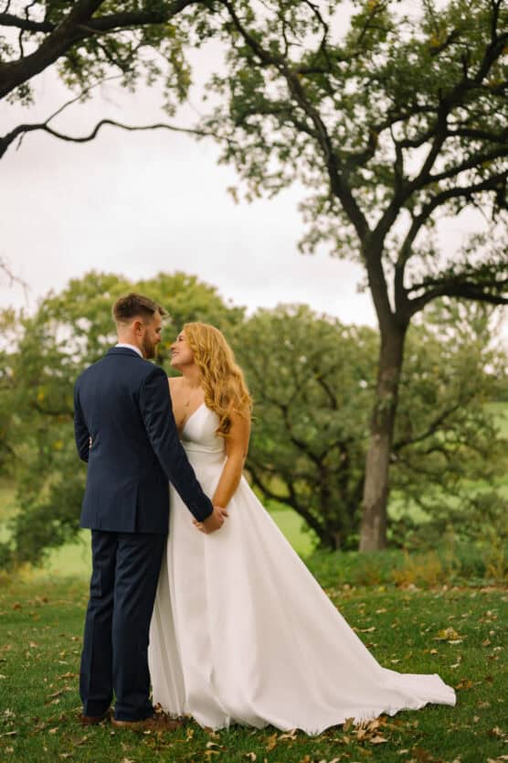 A bride in a white gown and groom in a navy suit stand outdoors on grass, holding hands and gazing at each other, surrounded by green trees under a cloudy sky. by Justin Salem Meyer