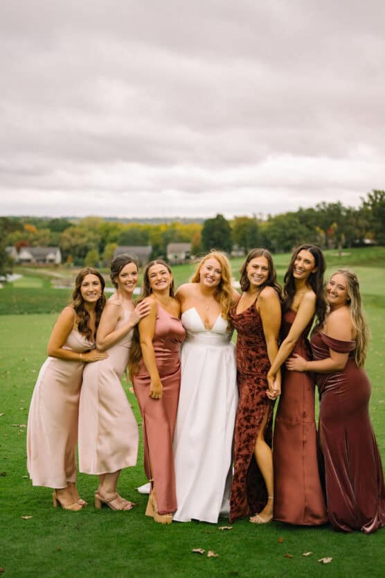 Seven women stand together on a grassy field. The woman in the center wears a white dress, and the others wear dresses in shades of pink, rose, and burgundy. They are smiling and posing closely for the photo. by Justin Salem Meyer