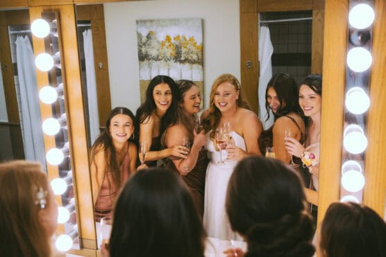 Six women, including a bride in a white dress, smile and hold champagne glasses while standing together in front of a mirror surrounded by bright lights in a dressing room. by Justin Salem Meyer